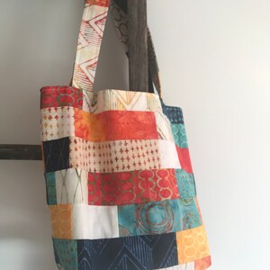 K&ouml;nnte beinhalten: Eine Patchwork-Tragetasche mit einem farbenfrohen Design aus orangefarbenem, rotem, blauem, gelbem und wei&szlig;em Stoff. Die Tasche hat einen einzigen Trageriemen und h&auml;ngt an einer Holzleiter.
