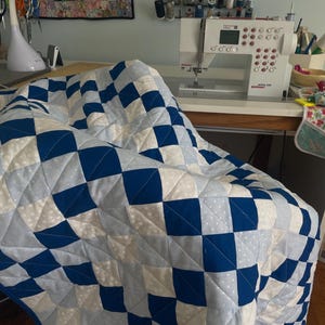 May include: A blue and white patchwork quilt with a checkerboard design. The quilt is draped over a chair. A sewing machine and sewing accessories are visible in the background, suggesting a craft or hobby.
