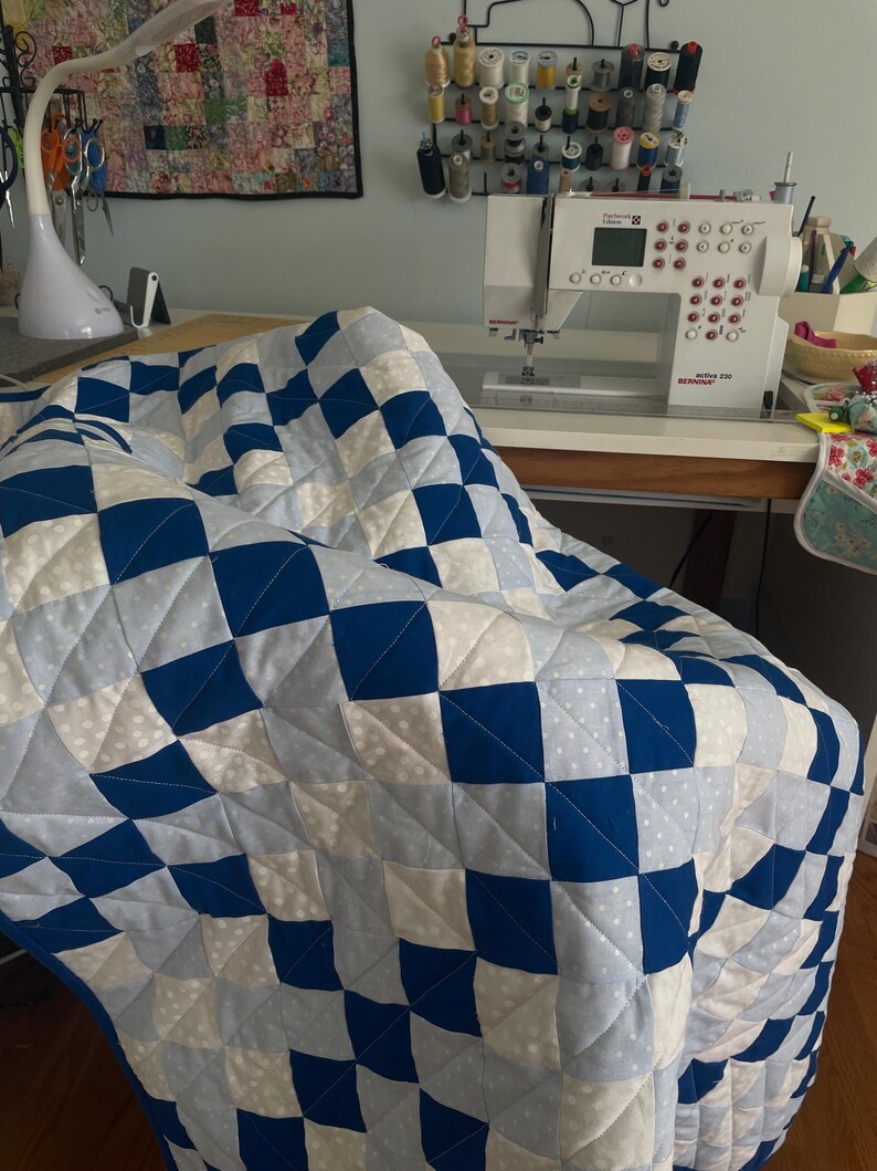 May include: A blue and white patchwork quilt with a checkerboard design. The quilt is draped over a chair. A sewing machine and sewing accessories are visible in the background, suggesting a craft or hobby.