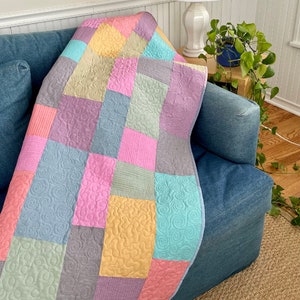 Op de afbeelding: Een patchworkdeken met verschillende pastelkleuren en draaiende patronen. De deken is gemaakt van vierkanten en rechthoeken, en de kleuren omvatten roze, blauw, groen, geel en paars.