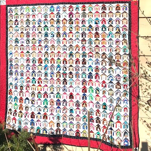 Pode incluir: Um edredom grande e colorido com um padrão repetido de casinhas de cachorro, cada uma com uma raça de cachorro diferente dentro. O edredom tem um fundo branco com uma borda vermelha e acabamento preto. As casinhas são de várias cores.