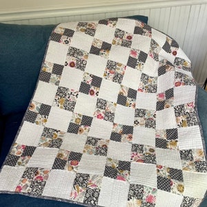 Peut inclure: Une courtepointe à carreaux avec un fond blanc et des carrés à motifs noirs, gris et floraux. La courtepointe est pliée et posée sur un canapé bleu.