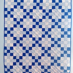 May include: A square quilt featuring a blue border and a repeating pattern of blue and white squares. The quilt is machine-stitched with a grid-like design, creating a visually appealing geometric pattern. The colour scheme is a classic combination of blue and white.