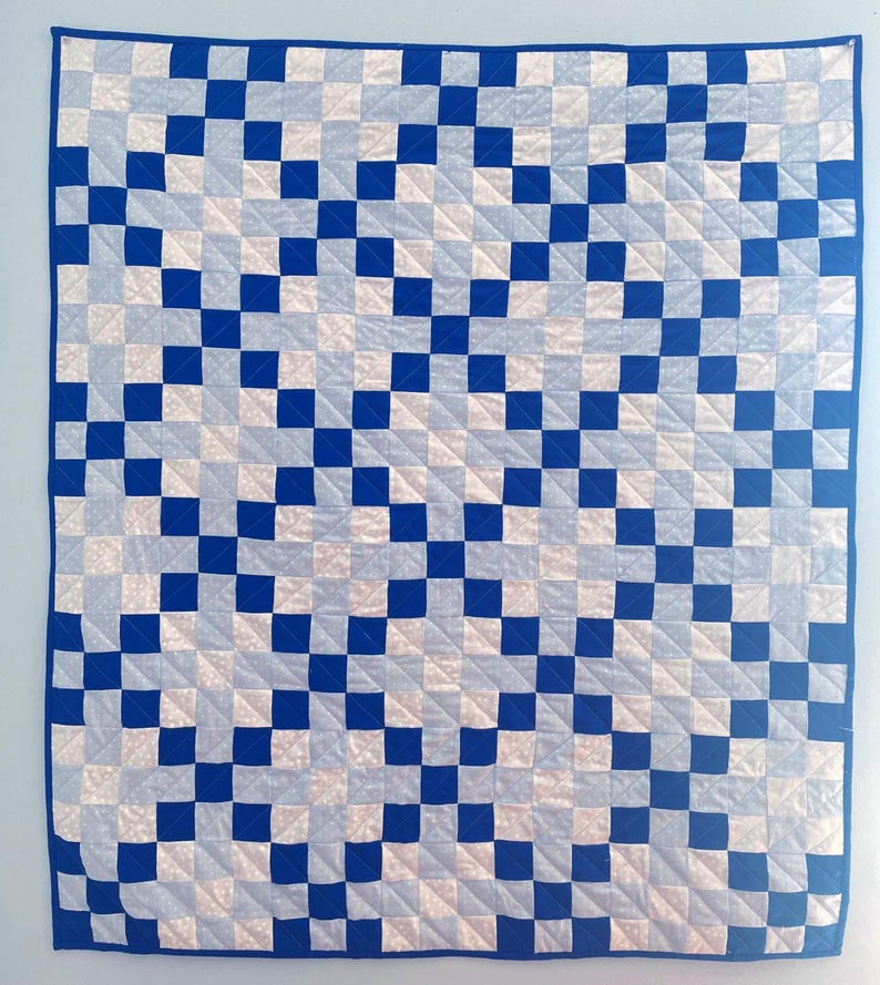 May include: A square quilt featuring a blue border and a repeating pattern of blue and white squares. The quilt is machine-stitched with a grid-like design, creating a visually appealing geometric pattern. The colour scheme is a classic combination of blue and white.