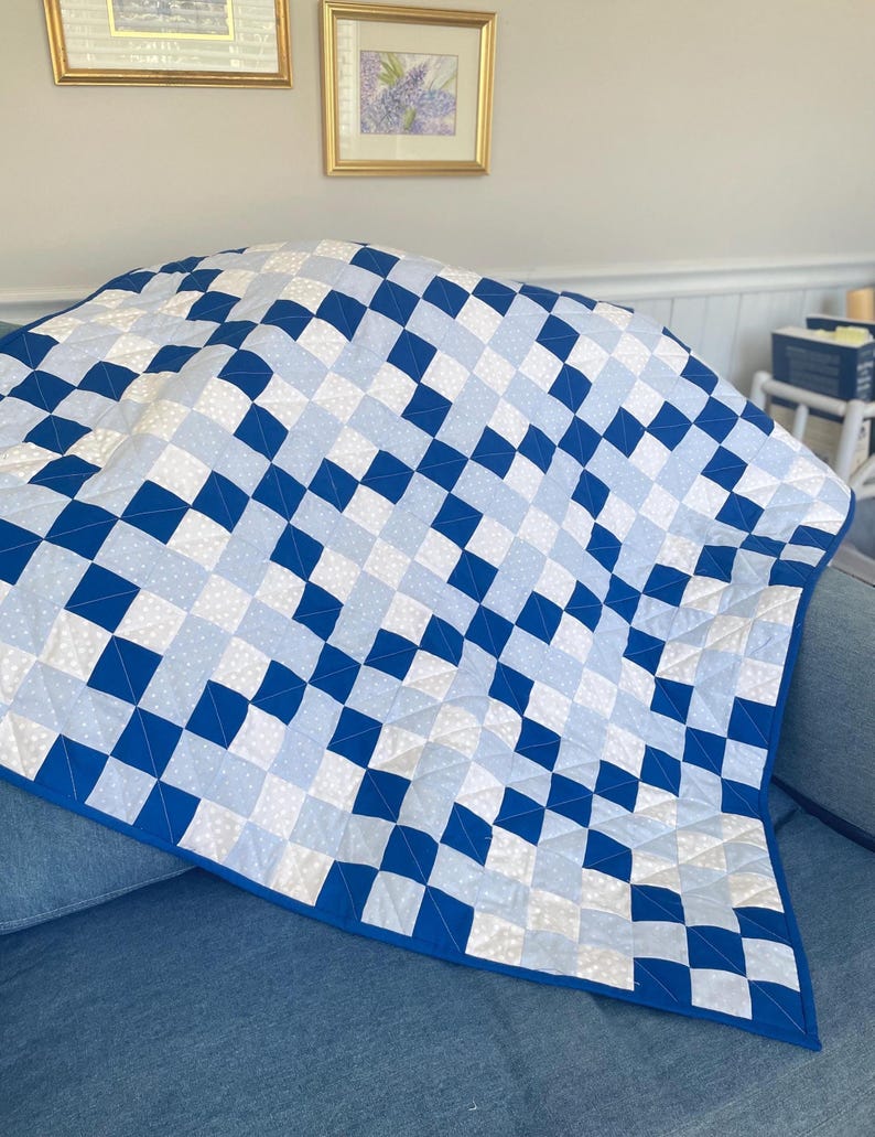 May include: A blue and white quilt with a geometric pattern of squares and diamonds. The quilt has a solid blue border and is draped over a blue surface. The quilt is a handmade item.