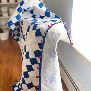 May include: A handmade quilt featuring a blue and white chequered design. The edges are secured with red and clear clips. The quilt is draped over a surface, highlighting the intricate stitching and overall pattern.