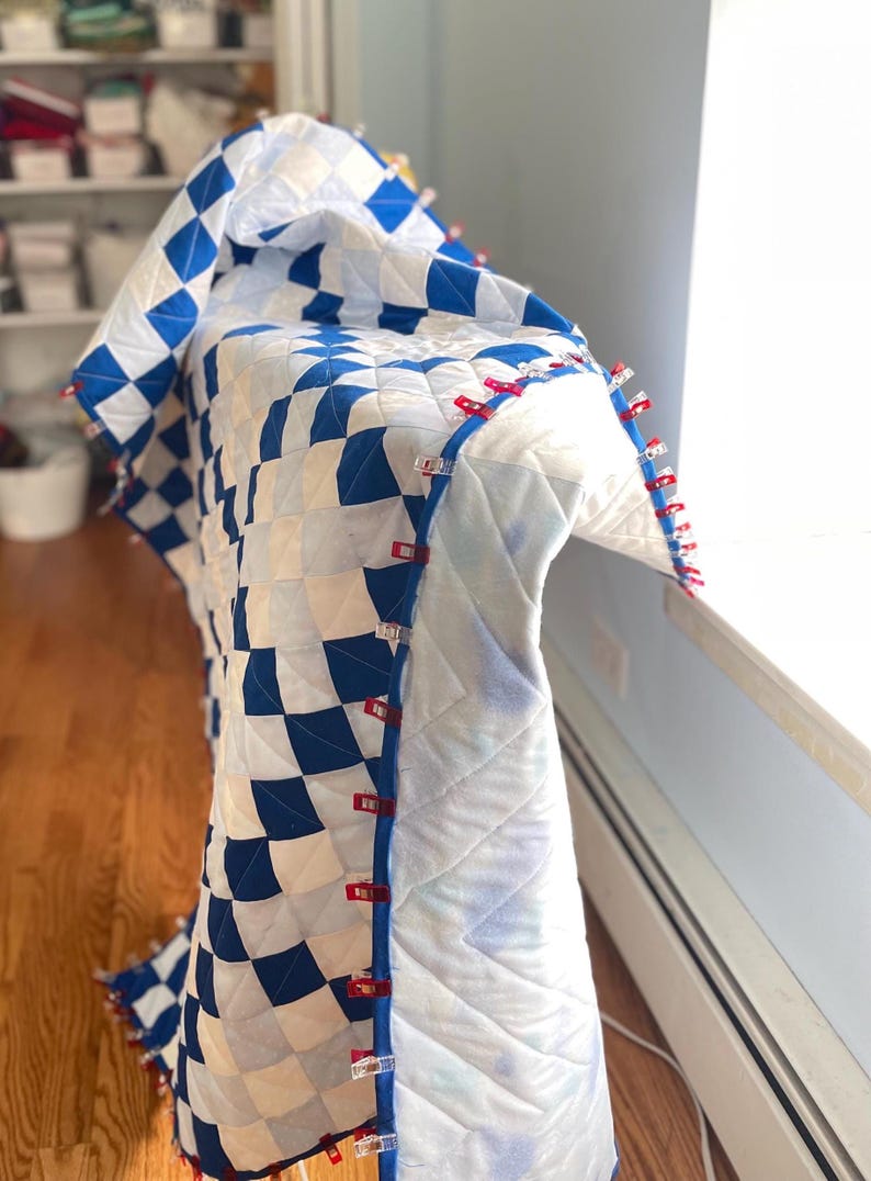 May include: A handmade quilt featuring a blue and white chequered design. The edges are secured with red and clear clips. The quilt is draped over a surface, highlighting the intricate stitching and overall pattern.