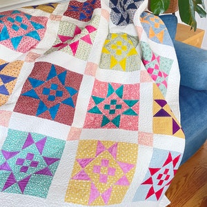 May include: A colorful patchwork quilt with a variety of star patterns in shades of pink, yellow, blue, green, and purple. The quilt is made of different fabrics and textures, and is folded on a blue couch.