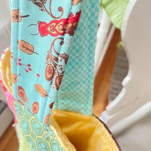 K&ouml;nnte beinhalten: Eine Patchwork-Stofftasche mit gelbem Futter und einem blau-wei&szlig; karierten Trageriemen. Die Tasche hat ein Blumenmuster auf der Vorderseite.