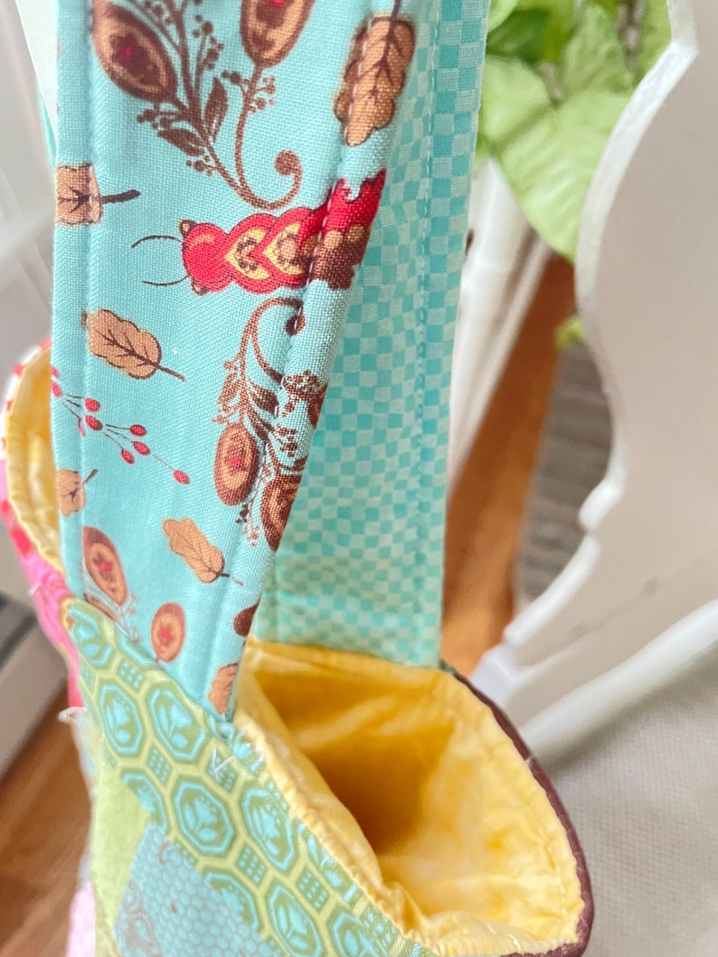K&ouml;nnte beinhalten: Eine Patchwork-Stofftasche mit gelbem Futter und einem blau-wei&szlig; karierten Trageriemen. Die Tasche hat ein Blumenmuster auf der Vorderseite.