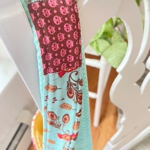 K&ouml;nnte beinhalten: Nahaufnahme einer blauen und braunen Stofftasche mit Patchwork-Muster. Die Tasche hat einen Riemen mit einem Muster aus rosa Marienk&auml;fern auf braunem Hintergrund.