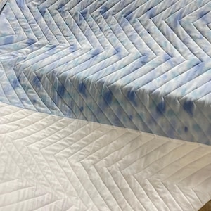 May include: Quilted fabric with a chevron design. The top section has a tie-dye effect in blue and white. The bottom section is plain white, creating a contrast. The fabric is likely for bedding or upholstery, with a modern aesthetic.