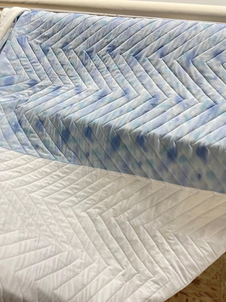 May include: Quilted fabric with a chevron design. The top section has a tie-dye effect in blue and white. The bottom section is plain white, creating a contrast. The fabric is likely for bedding or upholstery, with a modern aesthetic.