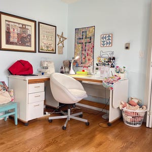 May include: A bright sewing room with a white desk, chair, and sewing machine. The room features framed artwork, a red bag, and a basket of fabric. The walls are painted a light blue, and the floor is hardwood.