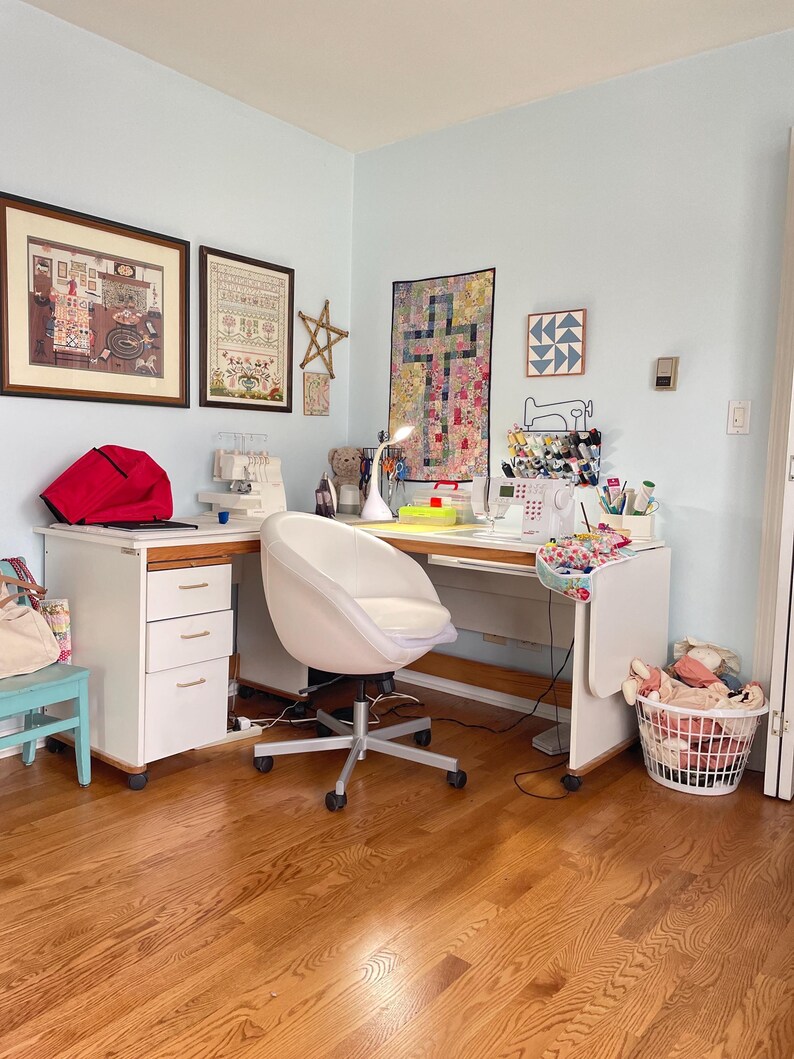 May include: A bright sewing room with a white desk, chair, and sewing machine. The room features framed artwork, a red bag, and a basket of fabric. The walls are painted a light blue, and the floor is hardwood.