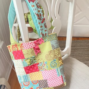 K&ouml;nnte beinhalten: Eine Patchwork-Tragetasche mit einem hellblauen Trageriemen. Die Tasche ist aus verschiedenen bunten Stoffen mit floralen und geometrischen Mustern gefertigt.