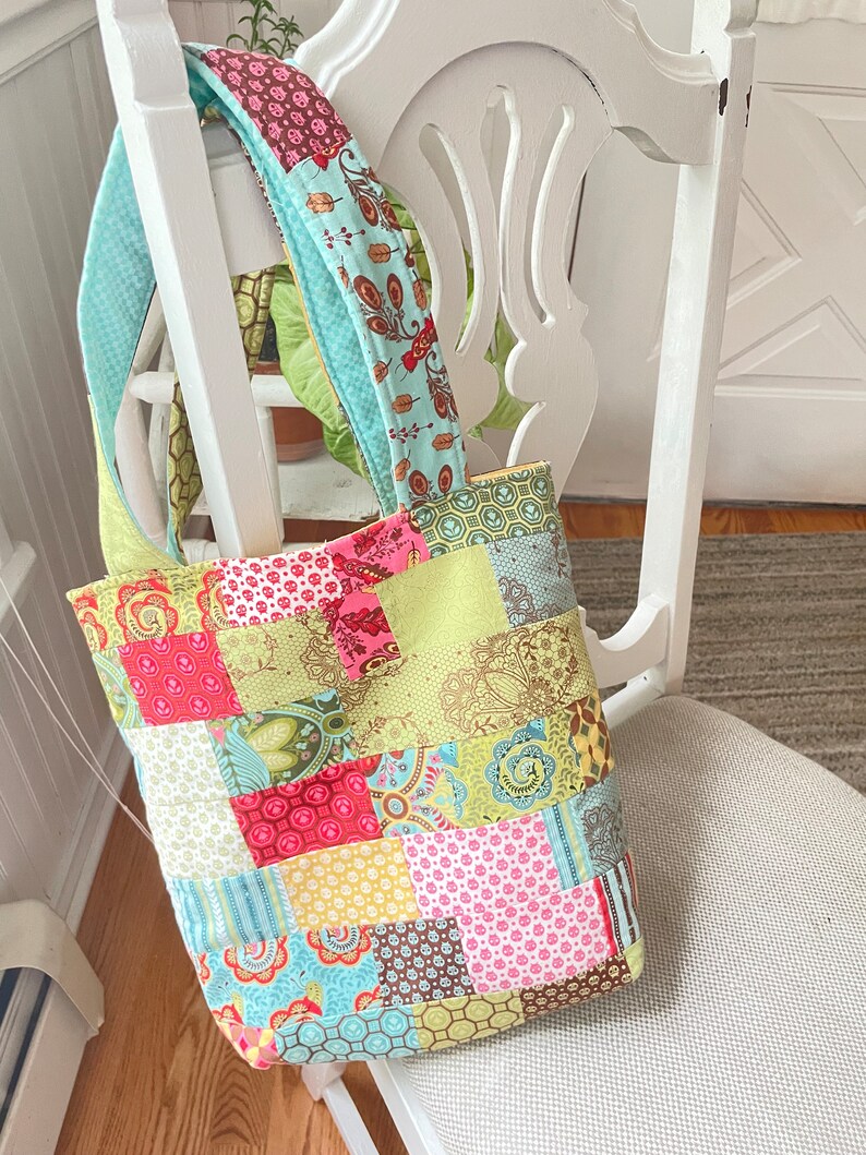 K&ouml;nnte beinhalten: Eine Patchwork-Tragetasche mit einem hellblauen Trageriemen. Die Tasche ist aus verschiedenen bunten Stoffen mit floralen und geometrischen Mustern gefertigt.