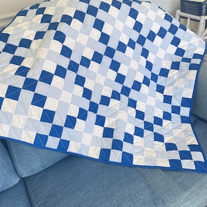May include: A blue and white quilt with a geometric pattern of squares. The quilt is draped over a blue sofa. The quilt's design features alternating squares of solid blue, light blue, and white with small polka dots.