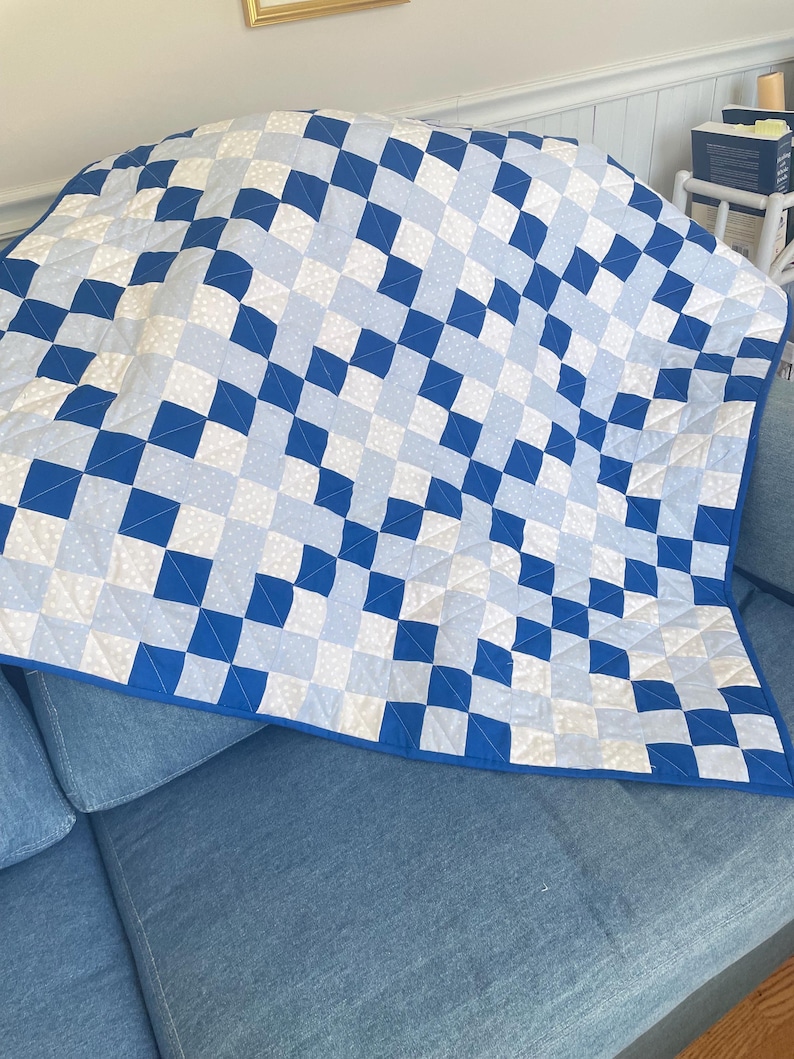 May include: A blue and white quilt with a geometric pattern of squares. The quilt is draped over a blue sofa. The quilt's design features alternating squares of solid blue, light blue, and white with small polka dots.