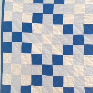 May include: A handmade quilt with a checkerboard pattern in shades of blue, white, and light grey. The squares are sewn together with a blue border. The quilt is made of soft fabric and is suitable for a baby or child.