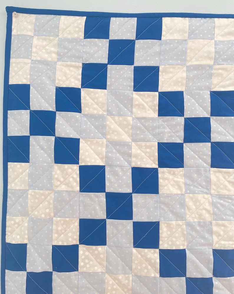 May include: A handmade quilt with a checkerboard pattern in shades of blue, white, and light grey. The squares are sewn together with a blue border. The quilt is made of soft fabric and is suitable for a baby or child.