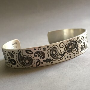 May include: A silver bracelet with an engraved paisley pattern. The bracelet is about 1/2 inch wide.