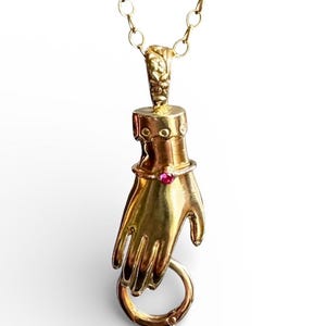 Puede incluir: Un collar con colgante dorado que representa una mano sosteniendo un anillo. La mano tiene una pequeña gema rosa en la muñeca. La cadena del collar también es dorada. El colgante es probablemente de metal.