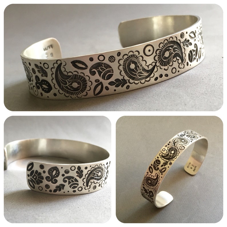 May include: A silver bracelet with a paisley pattern etched into the metal. The bracelet is open and has a slight curve to it.