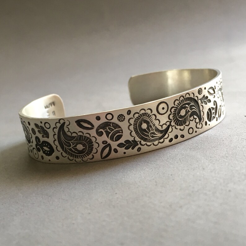 May include: A silver bracelet with an engraved paisley pattern. The bracelet is about 1/2 inch wide.