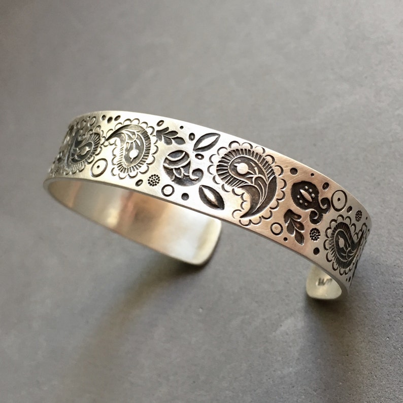 May include: A silver bracelet with a paisley design etched into the metal. The design is in a black color.