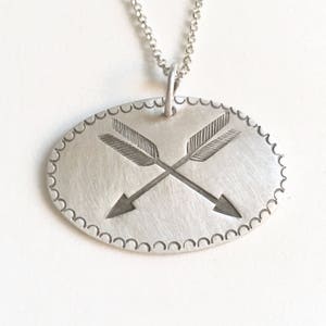 May include: A silver necklace with a pendant featuring two crossed arrows with feathered tips. The pendant is oval shaped and has a scalloped edge.