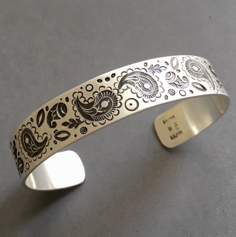 May include: A silver bracelet with a paisley pattern engraved on the front. The bracelet is stamped with the text "E.J.L.S."