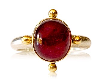 May include: A red gemstone ring with gold accents. The ring has a silver band and a round, cabochon-cut gemstone.