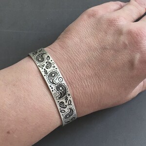 May include: A silver bracelet with a black paisley design. The bracelet is worn on a person's wrist.