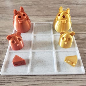 Può includere: Un tabellone da gioco bianco con una griglia di nove caselle. Ci sono quattro piccole figurine stampate in 3D, due dorate e due color rame, e due piccoli spicchi di formaggio stampati in 3D, uno dorato e uno color rame, sul tabellone.