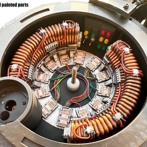 Può includere: Primo piano dell'interno di un modello, che mostra una bobina circolare di colore rame con fili rossi e neri. La bobina è circondata da una serie di piccoli pannelli rettangolari di colore argento con segni arancioni e neri. I pannelli sono disposti in un cerchio attorno alla bobina. L'interno è dipinto di colore grigio scuro.