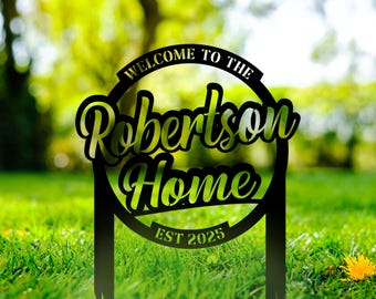Puede incluir: Letrero de jardín de metal negro con las palabras "Welcome to the Robertson Home" en una fuente decorativa. El letrero también incluye "EST 2025". El letrero está colocado en un jardín cubierto de hierba.