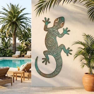 May include: A wall-mounted lizard sculpture featuring a weathered teal and bronze patina finish. The surface is decorated with intricate geometric patterns, including a central mandala design on the torso and spiral motifs along the curved tail.