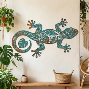 Puede incluir: Una gran pieza de arte mural de metal con forma de gecko, con un acabado desgastado en color turquesa y marrón. El gecko presenta intrincados diseños de hojas y espirales recortadas. El lagarto está montado en una pared blanca, con plantas y un banco de madera en primer plano.