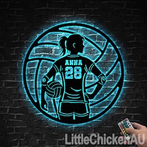 May include: A black metal volleyball player silhouette with the name "Anna" and the number "28" on the jersey, set within a volleyball outline. The design is illuminated with a blue neon glow, mounted on a dark brick wall.