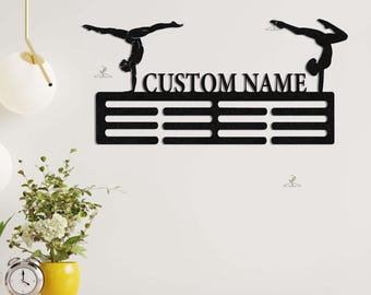 May include: Black metal medal display with two gymnast silhouettes. The display has 10 slots for medals and a space for a custom name. The name "CUSTOM NAME" is printed on the display.