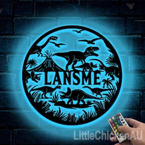 May include: A blue light illuminates a black metal cutout of a dinosaur scene with the name "LANSMEE" in the center. The scene includes a volcano, palm trees, and various dinosaurs. A remote control with multiple colored buttons is in the lower right corner.