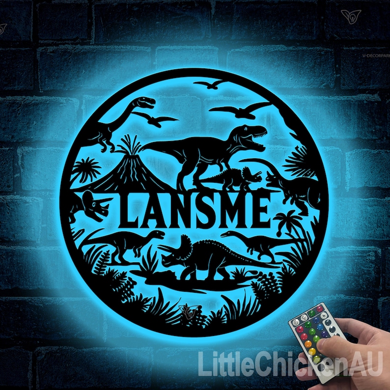 May include: A blue light illuminates a black metal cutout of a dinosaur scene with the name "LANSMEE" in the center. The scene includes a volcano, palm trees, and various dinosaurs. A remote control with multiple colored buttons is in the lower right corner.