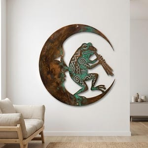 May include: A metal wall art piece depicting a frog playing a flute while perched on a crescent moon. The frog is green with intricate designs, and the moon has a face carved into it. The artwork has a rustic, bronze-like finish.