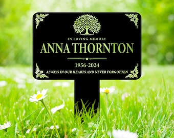 Puede incluir: Una estaca de jardín conmemorativa negra con un diseño de árbol y el texto "IN LOVING MEMORY ANNA THORNTON 1956-2024 ALWAYS IN OUR HEARTS AND NEVER FORGOTTEN". El letrero tiene detalles decorativos en las esquinas y está colocado en un área cubierta de hierba.