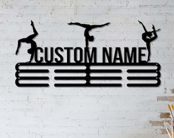 May include: Black metal gymnastics medal holder with three gymnast silhouettes and the text "CUSTOM NAME". The holder has three horizontal bars for displaying medals. The background is a white brick wall.
