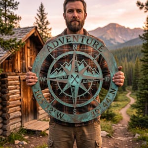 Pode incluir: Uma peça de arte de parede circular de metal com um design de bússola e a palavra "ADVENTURE" no topo. A bússola apresenta símbolos de montanha e direcionais, com as coordenadas 105° 12' W e 39° 44' N. A peça tem um acabamento turquesa envelhecido.