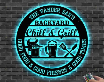 Könnte beinhalten: Schwarzes Metallschild mit dem Text "The Vander Sar's Backyard Chill & Grill", beleuchtet von blauem Neonlicht. Das Schild enthält Bilder eines Grills, eines Bierkrugs und gekreuzter Pfannenwender und Messer.