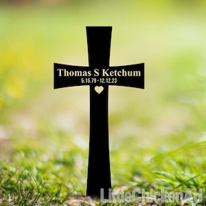 May include: A black cross-shaped memorial marker with the name "Thomas S Ketchum" and dates 5.15.79-12.12.23 in gold lettering. A small heart is below the dates. The background is blurred green grass.