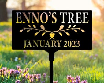 May include: Black metal garden sign with the text "ENNO'S TREE JANUARY 2023" in gold lettering. The sign features a decorative floral design above the date. The sign is placed in a garden with purple flowers.